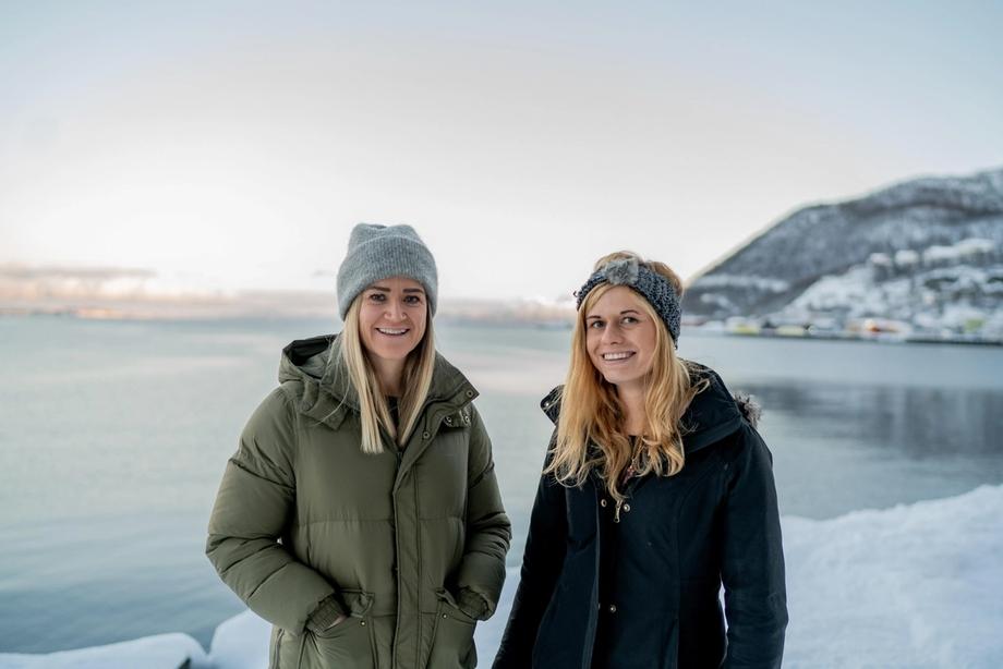 Portrettfoto av Linn Kristensen og Zoë Cumberpatch ved Equinor i Harstad som står utendørs i vinterklær, og med hav og snødekte fjell i bakgrunnen.