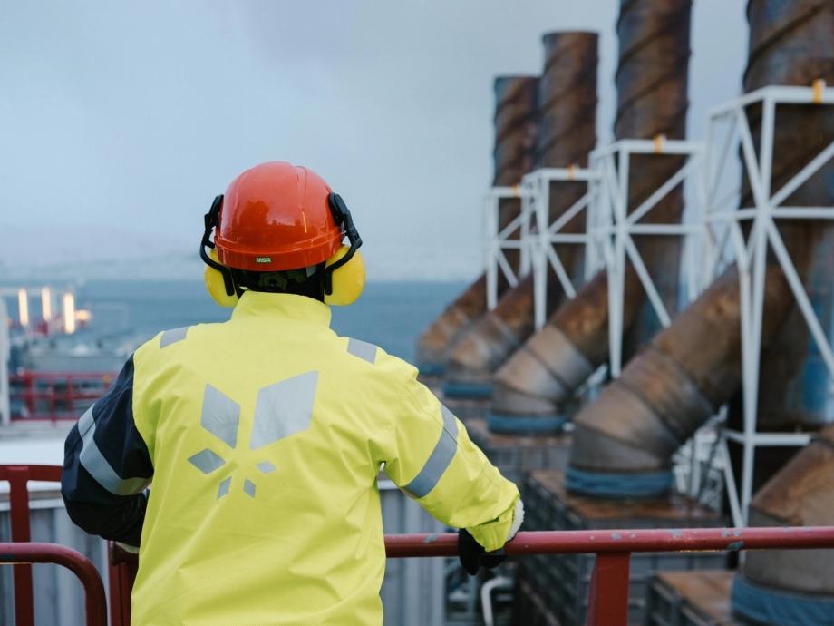 En person med rød vernehjelm og signalgul jakke med Equinor-logo står med ryggen til og ser utover gassanlegget på Melkøya. Havet skimtes i bakgrunnen.